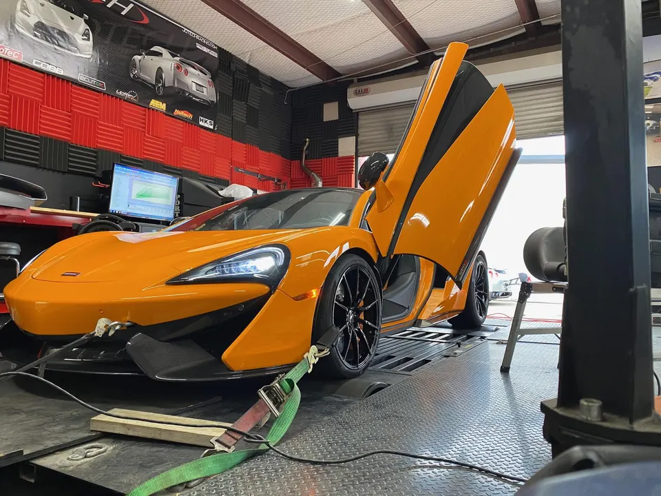Orange McLaren sports car with butterfly doors open, strapped to a dynamometer for performance tuning at a shop.
