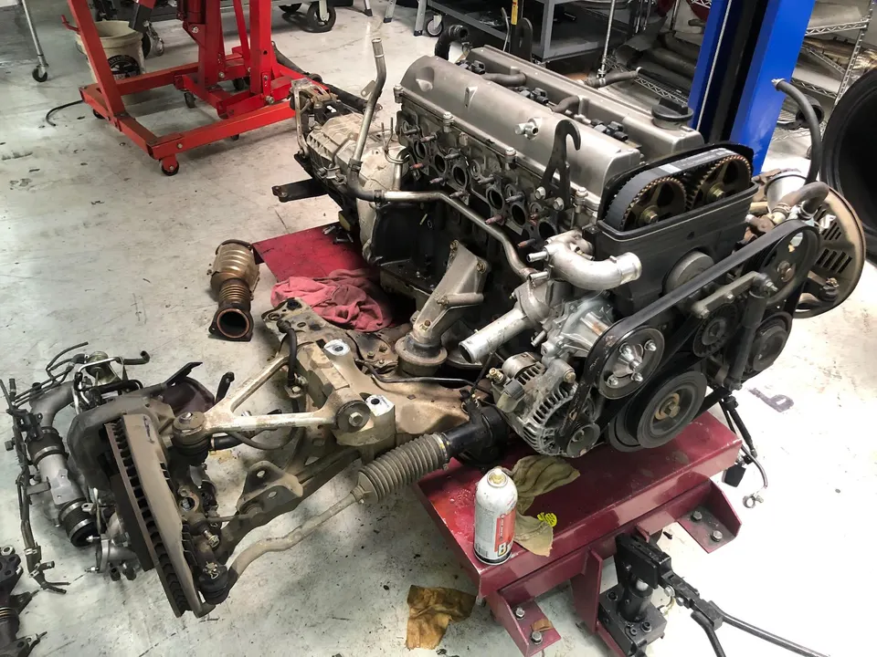 A disassembled 2JZ engine on a stand next to various suspension and drivetrain components on the floor of an automotive shop.