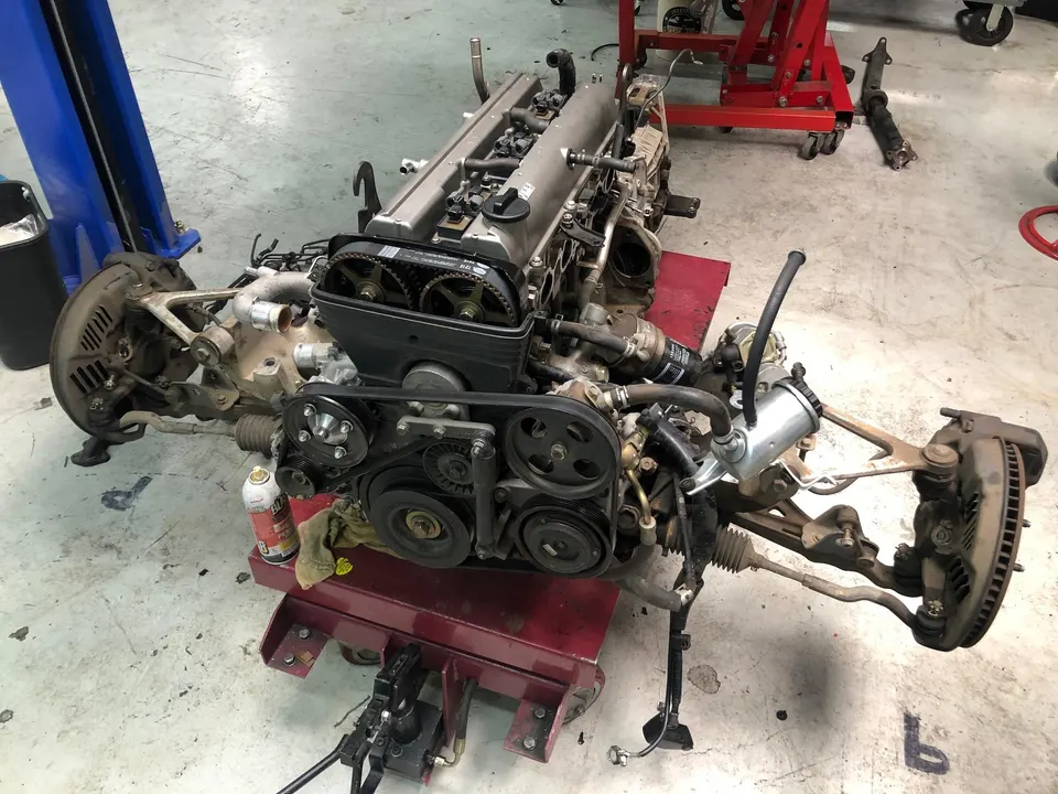 Toyota 2JZ engine and front suspension assembly on a stand in a shop, showing timing belt and pulleys.