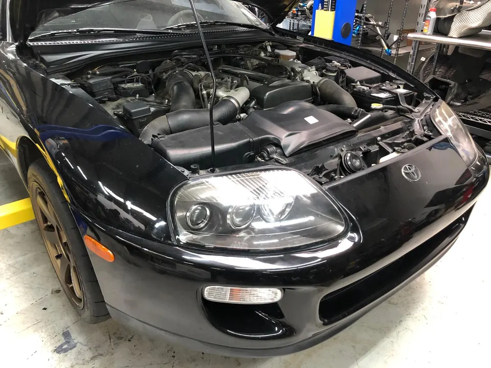 Black Toyota Supra with its hood open, revealing the engine bay and induction system in a shop setting.