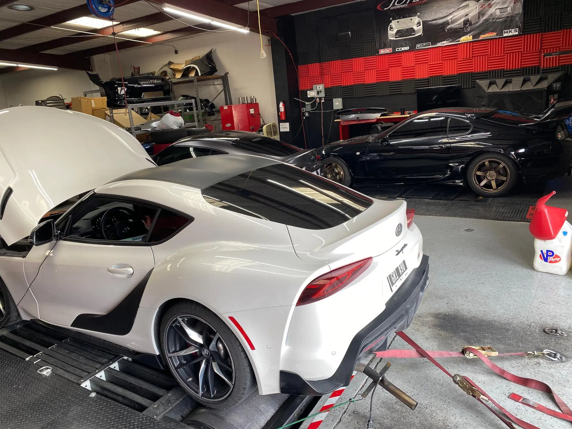 White Toyota Supra on a dyno with its hood open, being tuned at a performance shop, with a black Supra in the background.