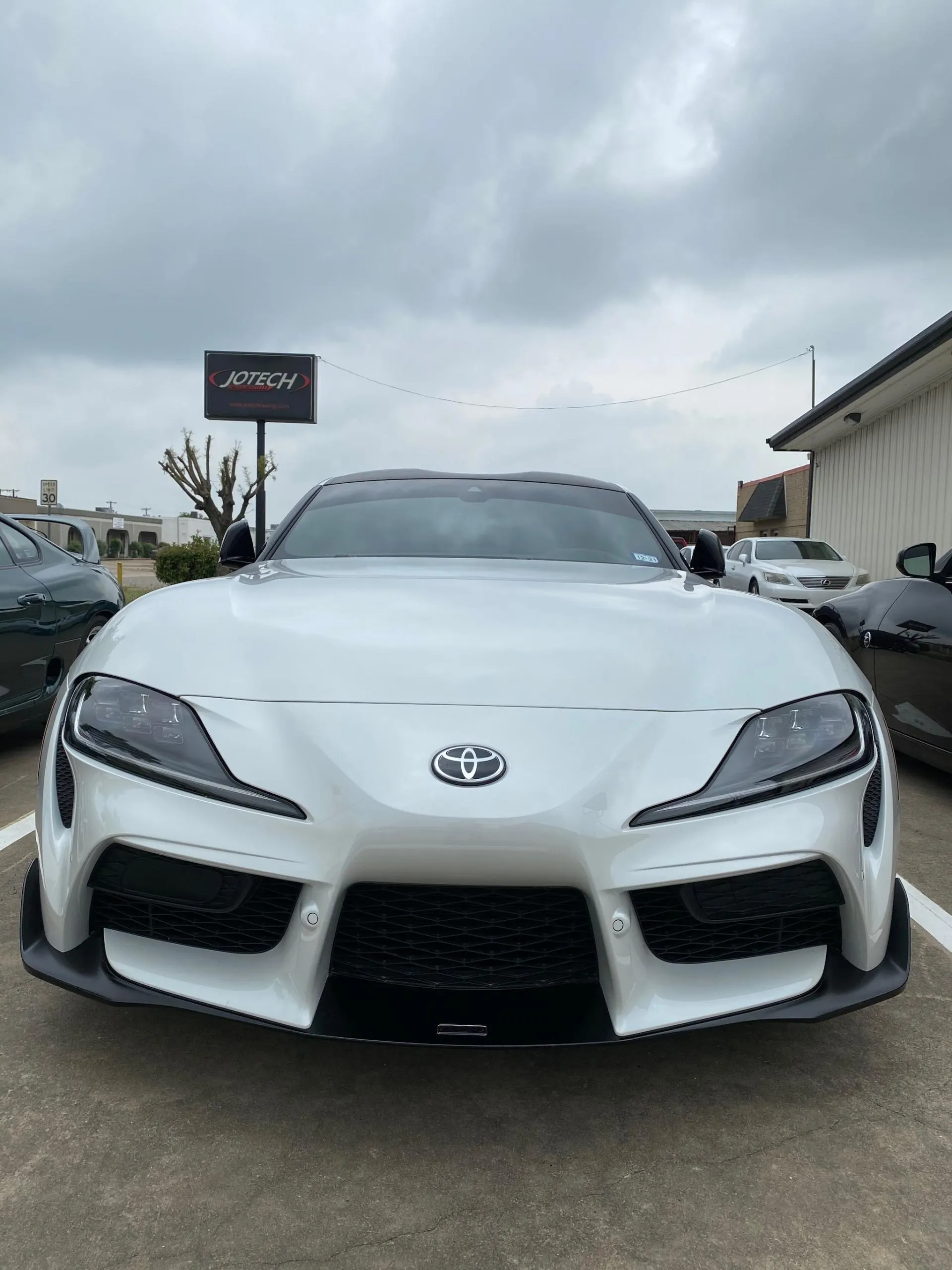Front view of a white Toyota Supra MK5 parked in a lot with a Jotech sign in the background.
