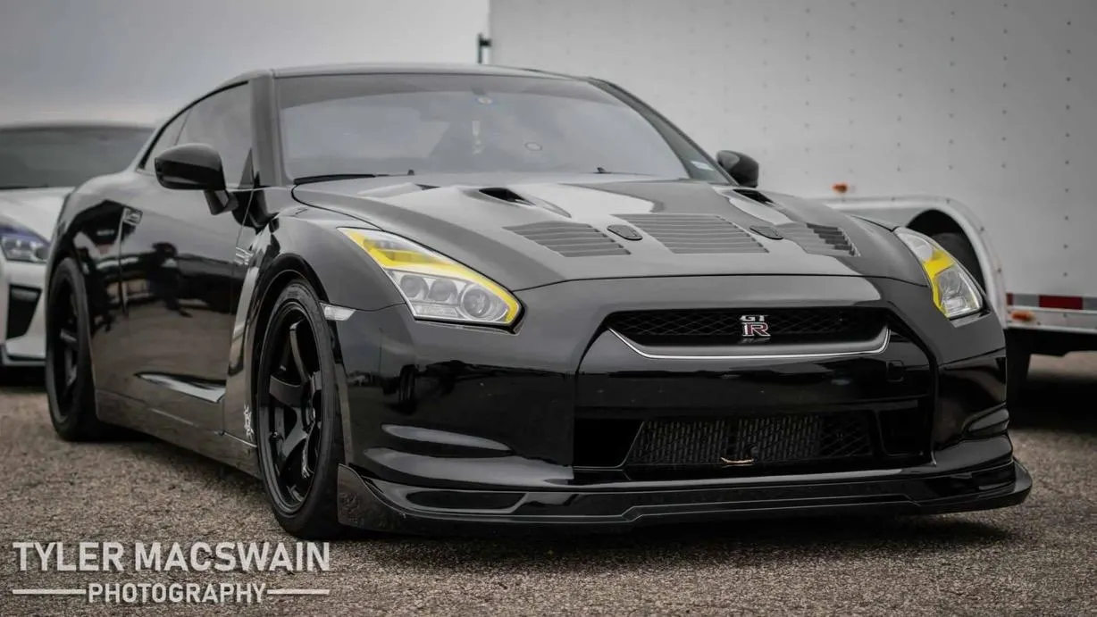Black Nissan GT-R with custom vented hood, front splitter, and yellow headlights parked outdoors.
