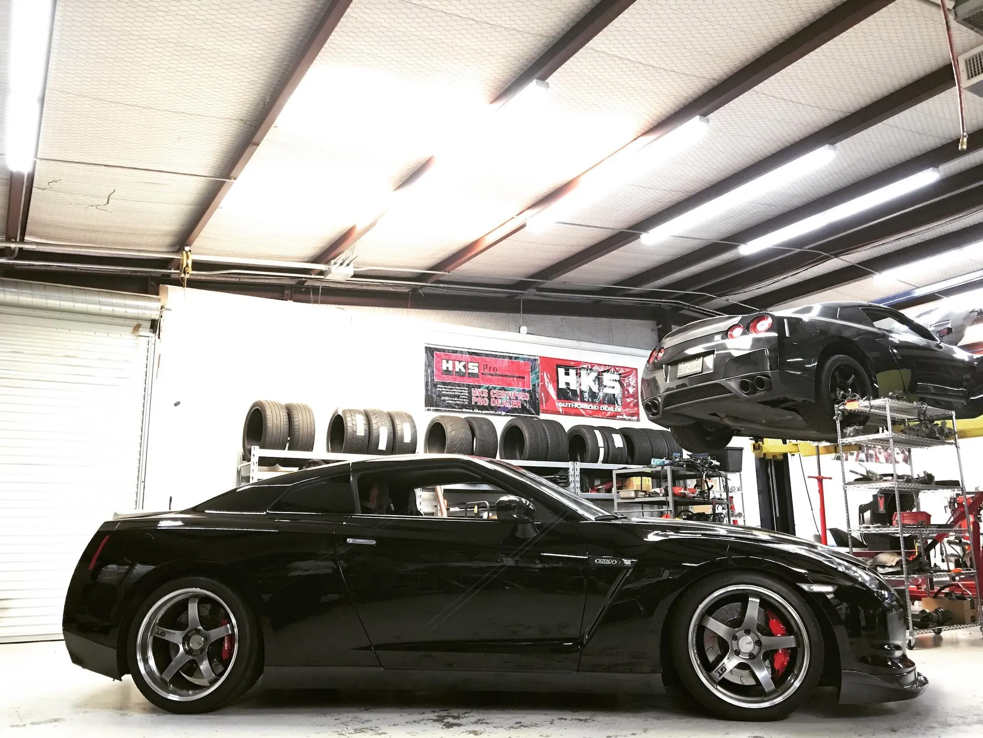 Two black Nissan GT-Rs in a performance tuning shop, one on a lift and one on the ground with aftermarket wheels.