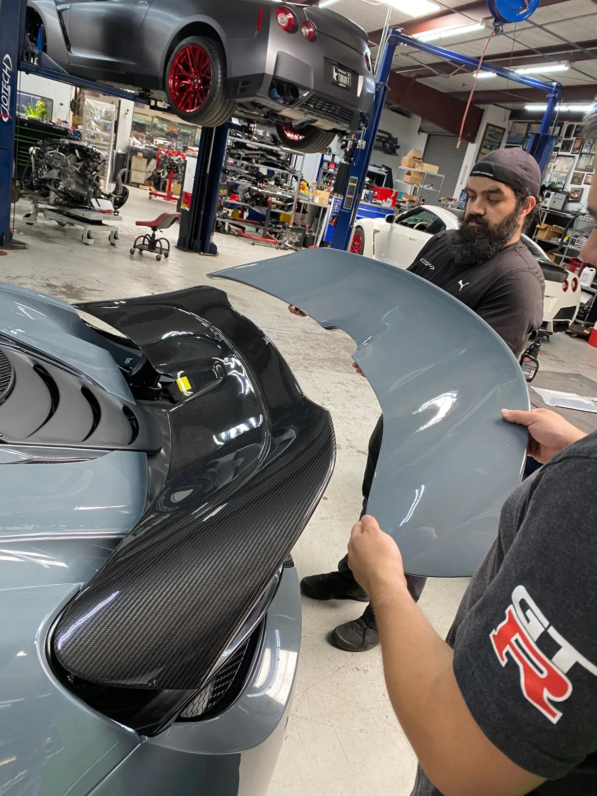 Two mechanics in a Jotech Motorsports shop comparing a grey spoiler to a carbon fiber wing on a McLaren, with a Nissan GT-R on a lift in the background.