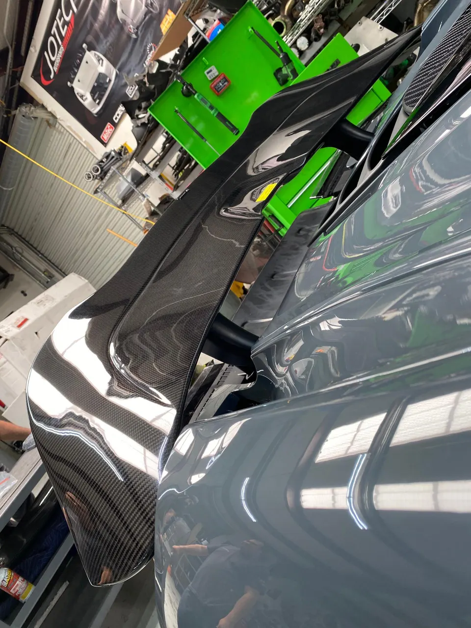 Close-up of a grey car's carbon fiber rear wing in a Jotech Motorsports shop.