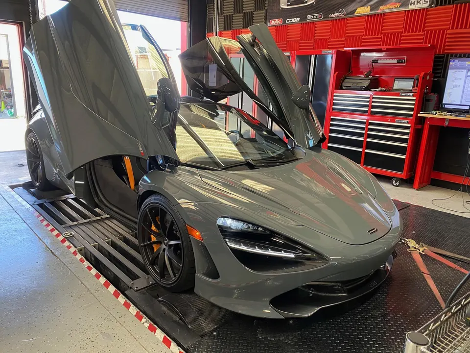 A grey McLaren 720S with its butterfly doors open, strapped to a dynamometer in a performance tuning shop.