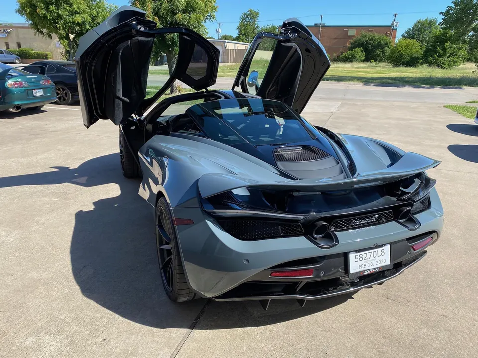Grey McLaren supercar with butterfly doors open, parked outdoors on a sunny day.