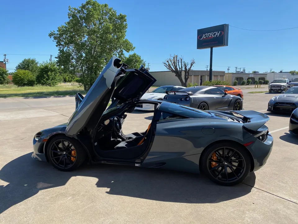 A grey McLaren 720S with its butterfly doors open, parked outside Jotech Motorsports with other performance cars.
