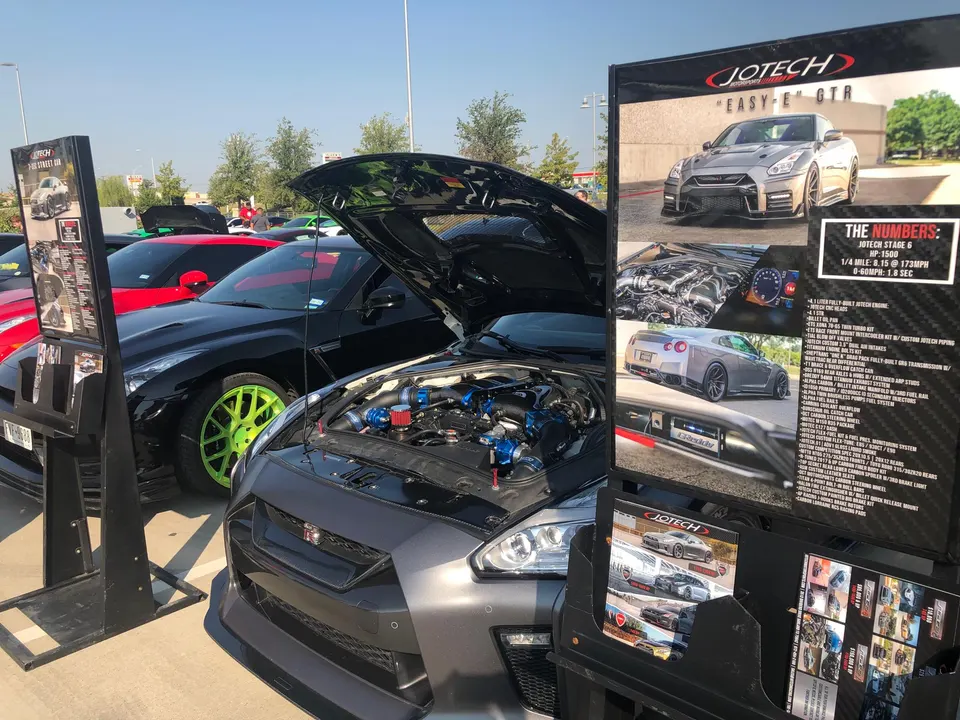 A grey Nissan GT-R with its hood open showcasing a modified engine bay, parked at an outdoor car show next to Jotech Motorsports display.