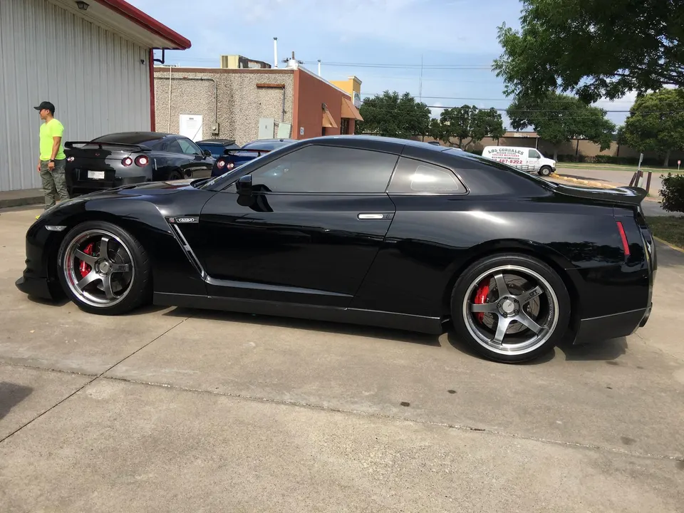 Black Nissan GT-R with aftermarket wheels and red brake calipers parked outdoors with other GT-Rs in the background.