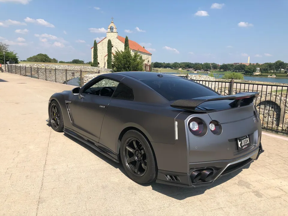 Matte grey Nissan GT-R with a carbon fiber wing and aftermarket wheels parked by a lake and church.