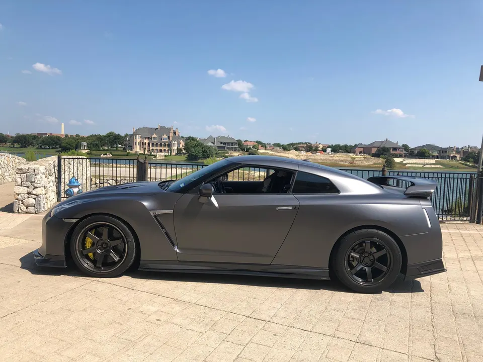 Matte grey Nissan GT-R R35 with aftermarket wheels and spoiler parked by a waterfront residential area on a sunny day.
