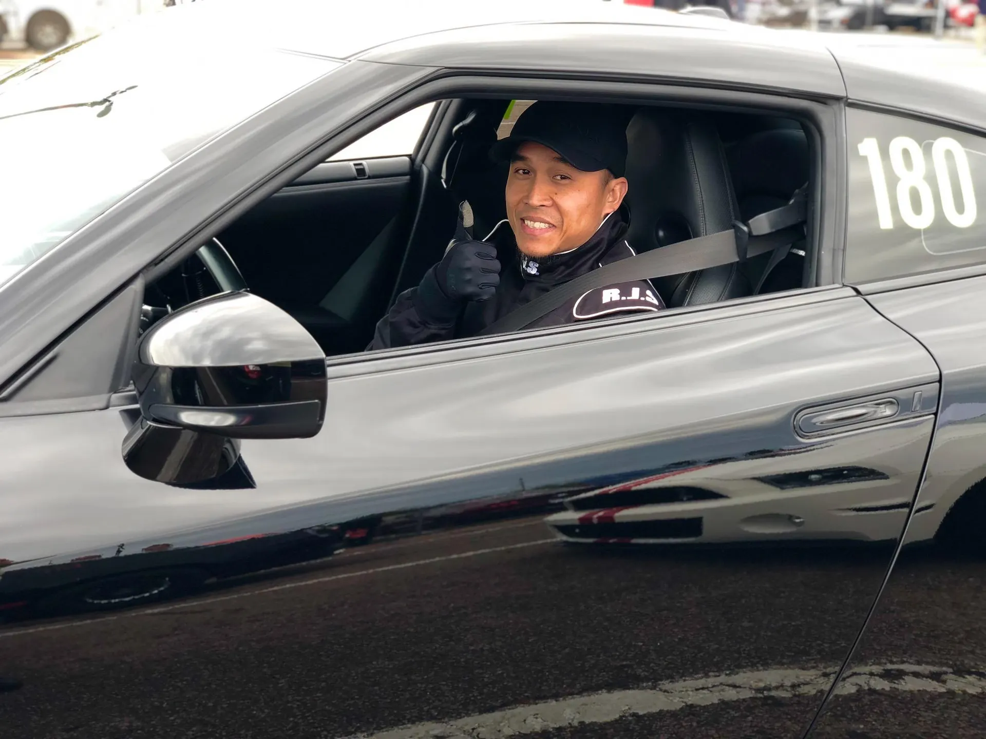 Driver in a black Nissan GT-R with race number 180 giving a thumbs up at a track event.