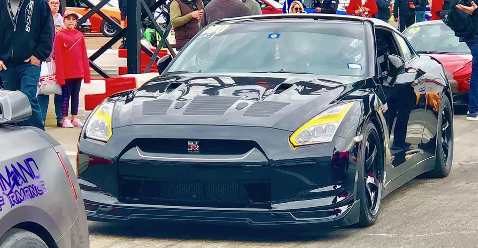 Black Nissan GT-R with yellow headlights and aftermarket wheels in a staging lane at a track event.