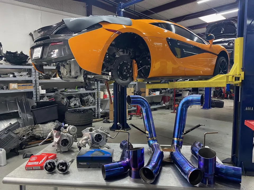 A McLaren supercar on a lift in a performance shop, with a custom blue exhaust system and turbochargers on a table.