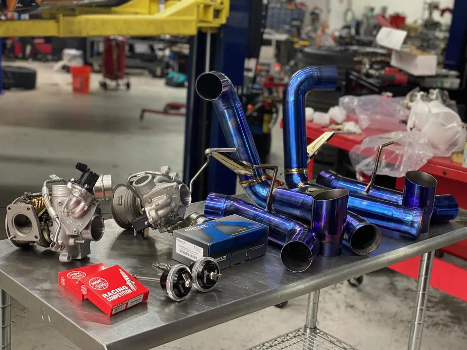 Performance car parts including turbochargers, titanium piping, spark plugs, and blow-off valves laid out on a table in a tuning shop.