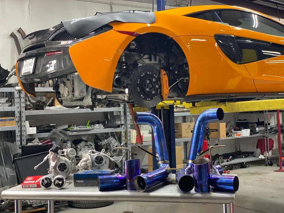 An orange McLaren on a lift in a performance shop, with a blue titanium exhaust system and turbos on a table.