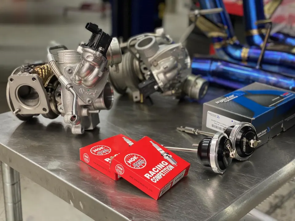 Performance car parts including turbochargers, wastegates, spark plugs, and injectors laid out on a shop bench.