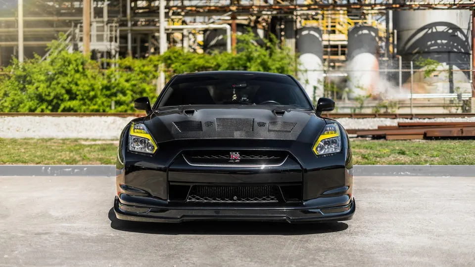A black Nissan GT-R with a carbon fiber hood and custom yellow headlights parked in front of an industrial facility.
