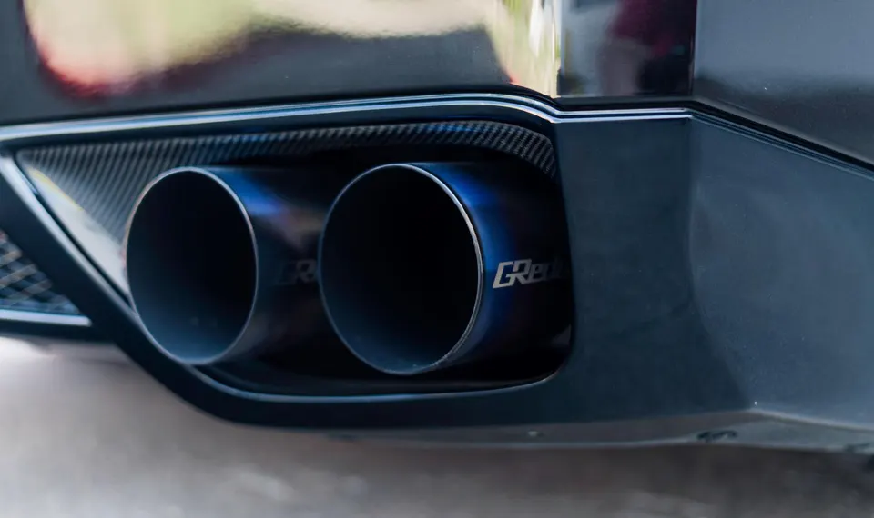 A close-up of a car's rear bumper showcasing GReddy aftermarket exhaust tips with blue finish and carbon fiber trim.