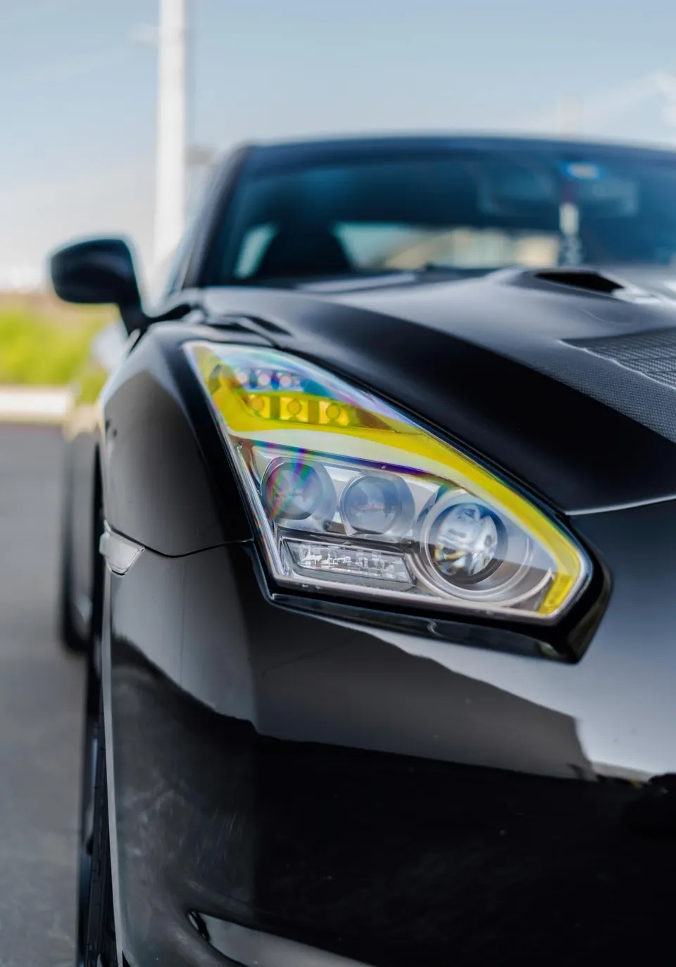 A close-up detail shot of the front headlight and fender of a black Nissan GT-R with yellow-tinted headlights.