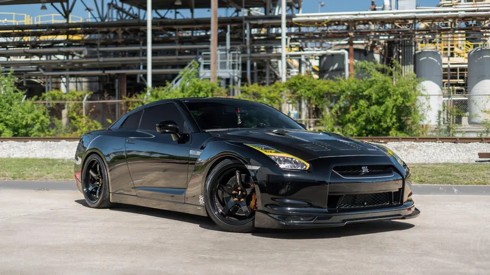 Black Nissan GT-R R35 with aftermarket wheels and yellow headlight accents parked in front of an industrial facility.