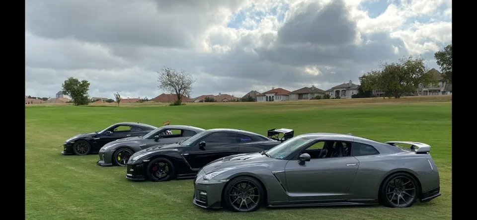 Four Nissan GT-R R35s, including grey and black models, parked in a line on a green golf course with houses in the background.