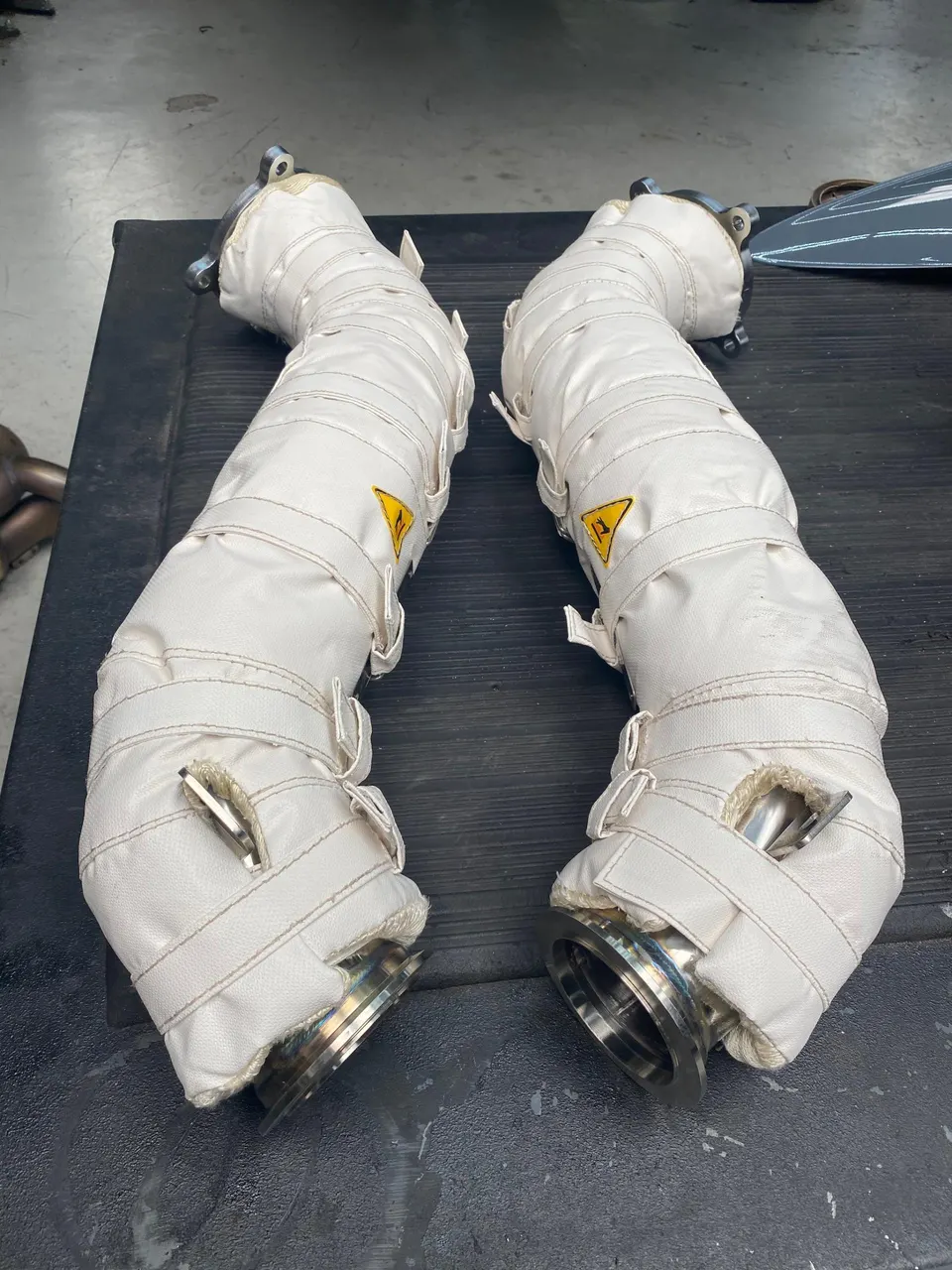 Two performance exhaust downpipes with white heat shielding on a workbench in a shop.