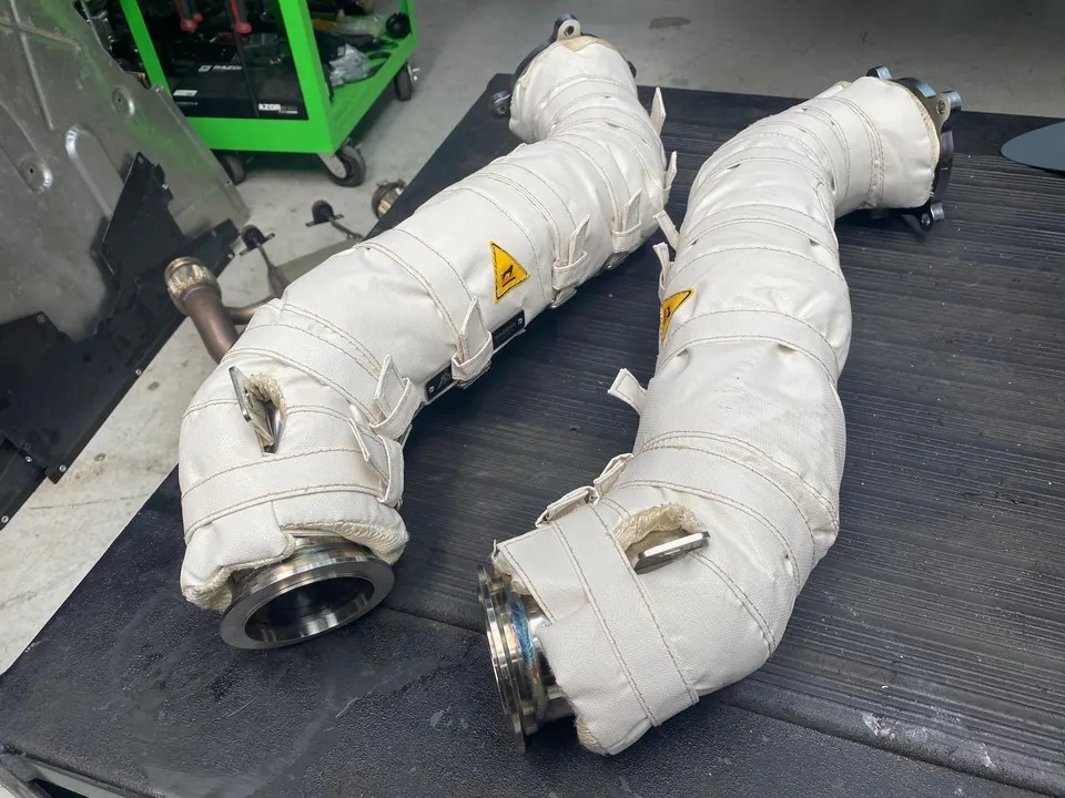 Two white heat-shielded exhaust downpipes with metal flanges on a black workbench in a performance shop.