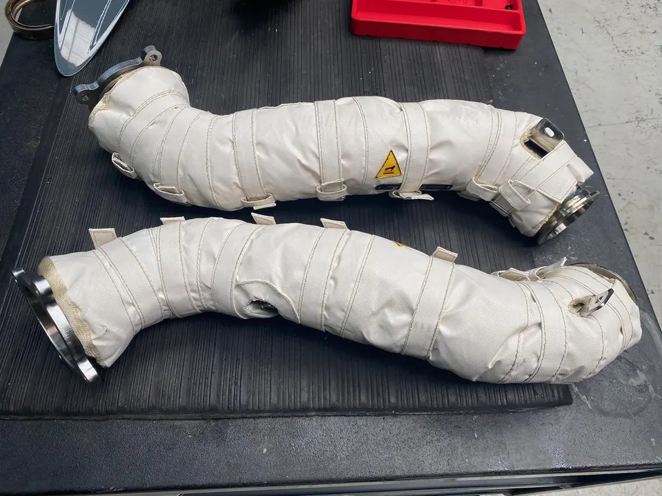 Two white heat-wrapped exhaust downpipes for a performance vehicle on a dark surface.