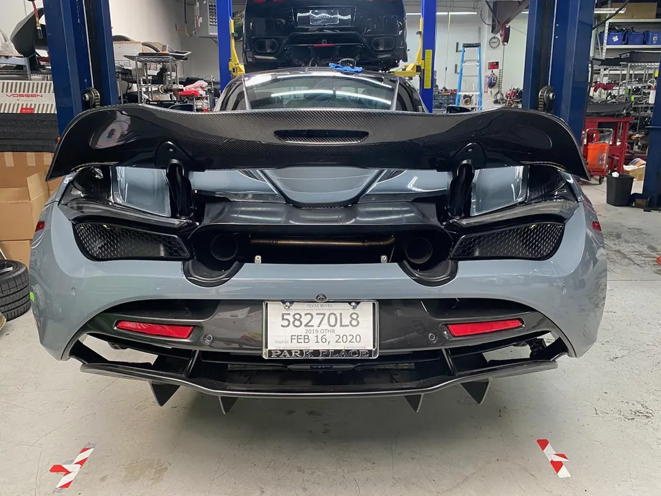 Rear view of a grey McLaren on a lift in a shop, showcasing its carbon fiber wing, exhaust, and diffuser.