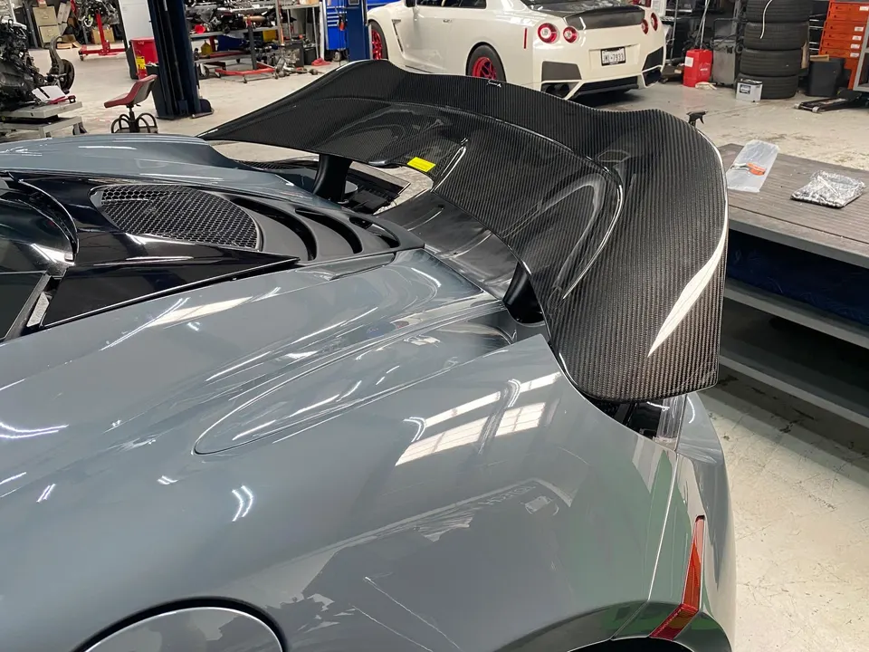 Rear view of a grey McLaren with a large carbon fiber wing in a performance tuning shop, with a white GT-R in the background.