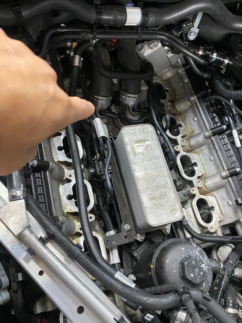 Mechanic's hand pointing at a car engine during disassembly, showing the cylinder head and oil cooler.