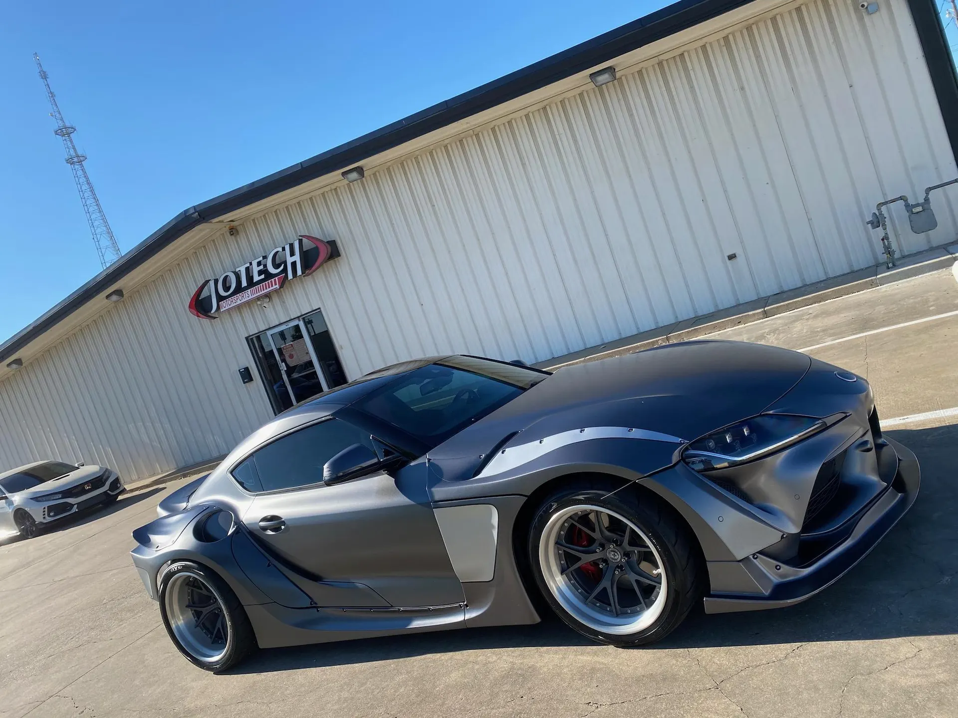 A matte grey widebody Toyota Supra A90 with custom wheels parked in front of the Jotech Motorsports shop.