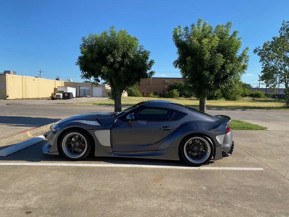 A grey widebody Toyota Supra with custom wheels parked outdoors on a sunny day, showcasing its aggressive exterior modifications.