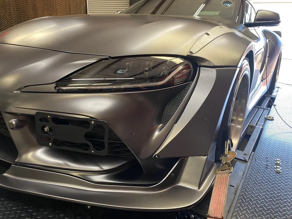 Close-up front view of a matte grey widebody Toyota Supra strapped down on a dyno for performance testing.