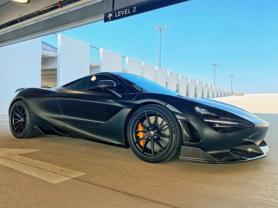 A black McLaren 720S supercar with carbon fiber aero and orange brake calipers parked in a modern parking structure.