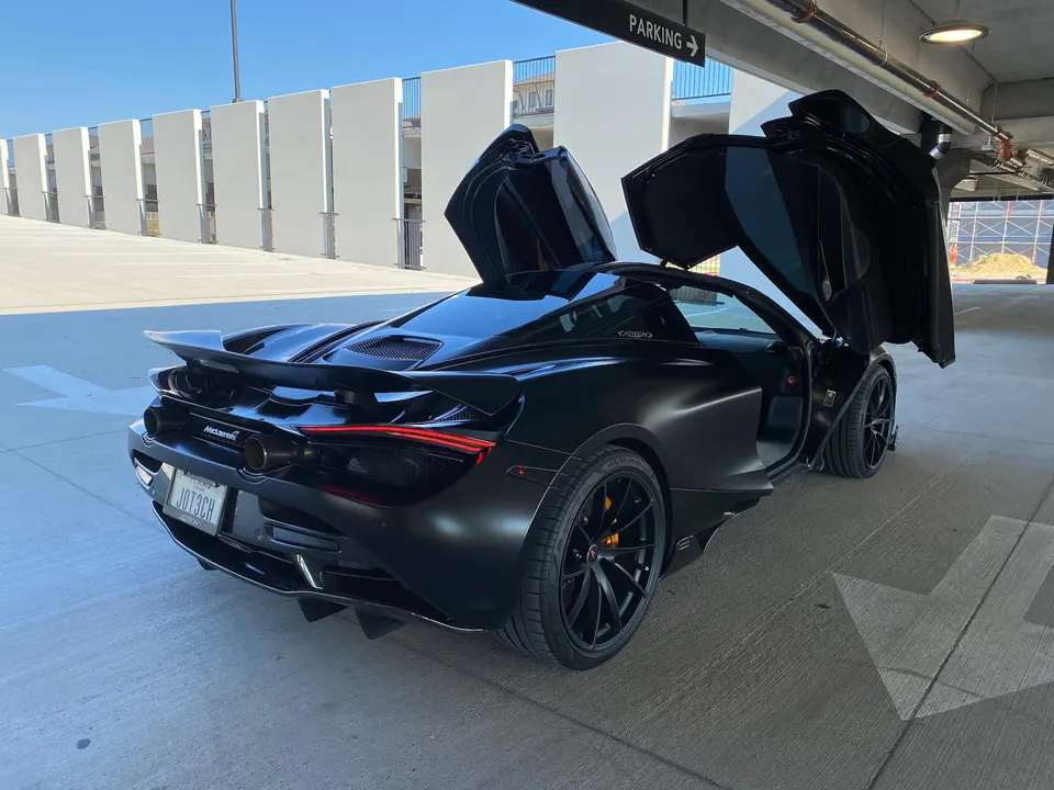 A black McLaren 720S with its butterfly doors open, parked in a bright parking garage.