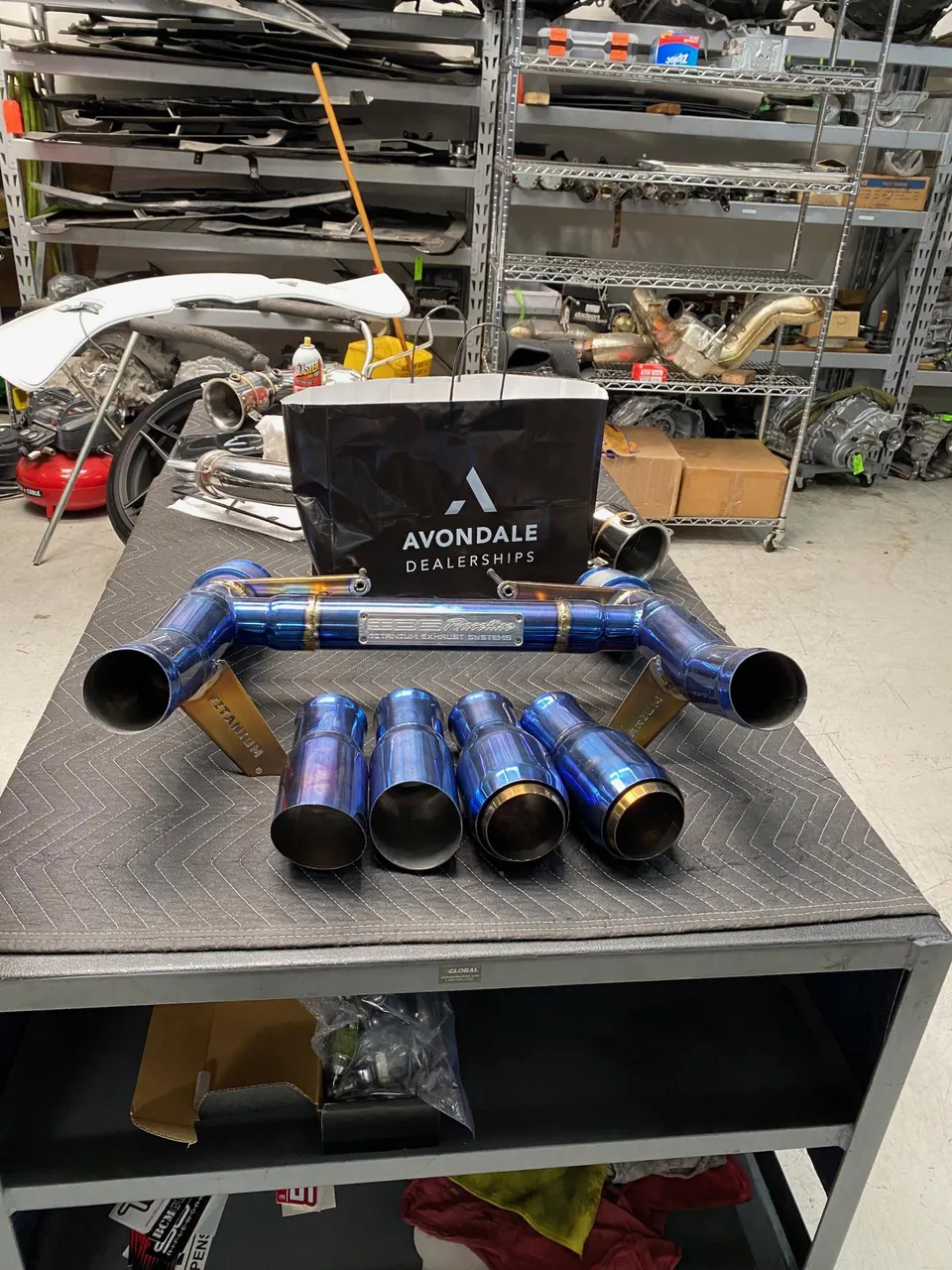 Titanium performance exhaust system with multiple tips displayed on a workbench in a car tuning shop, with an Avondale Dealerships bag.