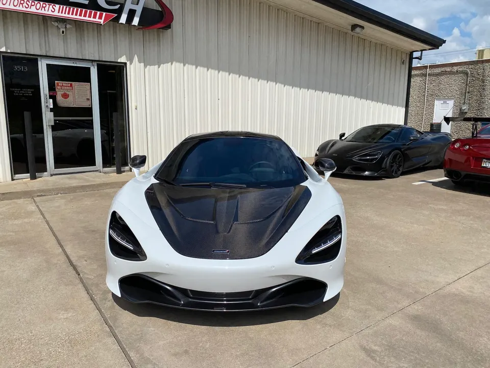 A white McLaren 720S with a carbon fiber hood parked outside Jotech Motorsports, with a black McLaren 720S and a red Nissan GT-R in the background.