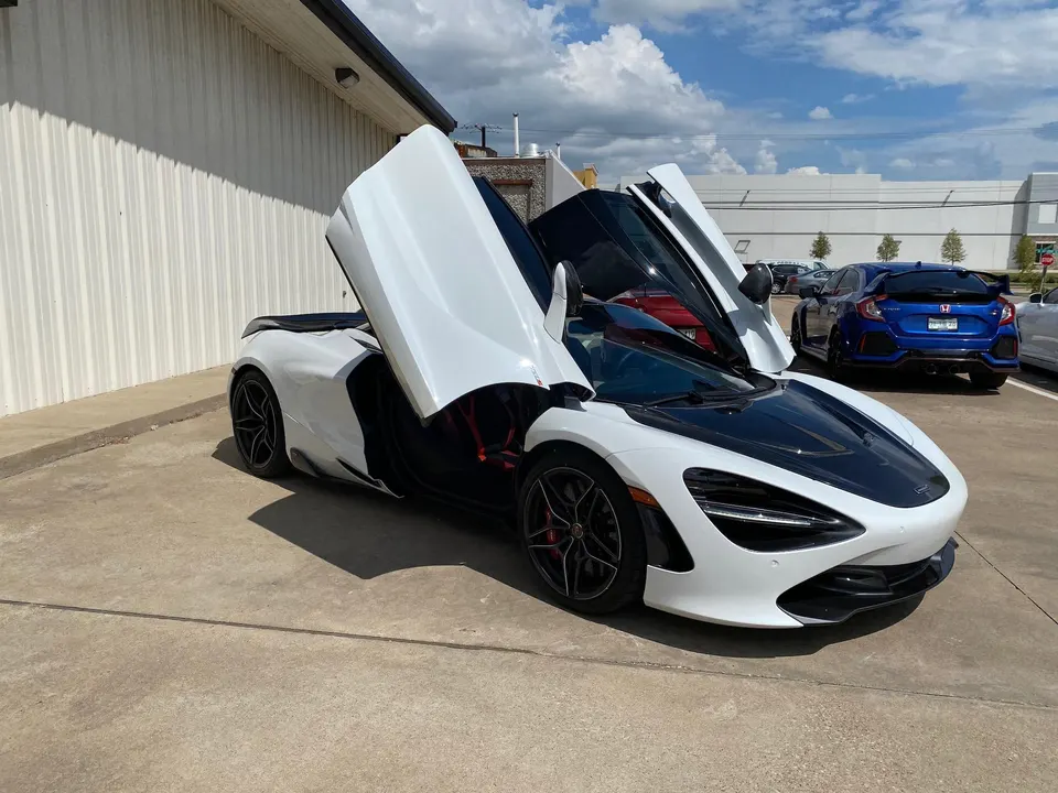 A white McLaren 720S with its butterfly doors open, parked in a lot on a sunny day.