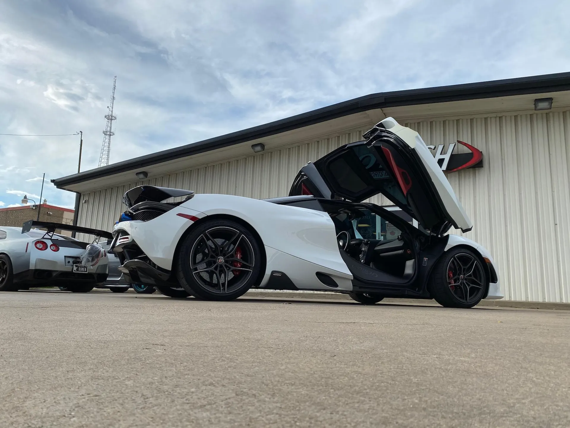 White McLaren 720S with open dihedral doors parked outside Jotech Motorsports, with a silver Nissan GT-R behind it.