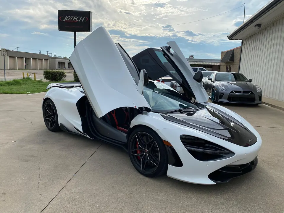 White McLaren 720S with butterfly doors open, parked outside Jotech Motorsports with a Nissan GT-R in the background.
