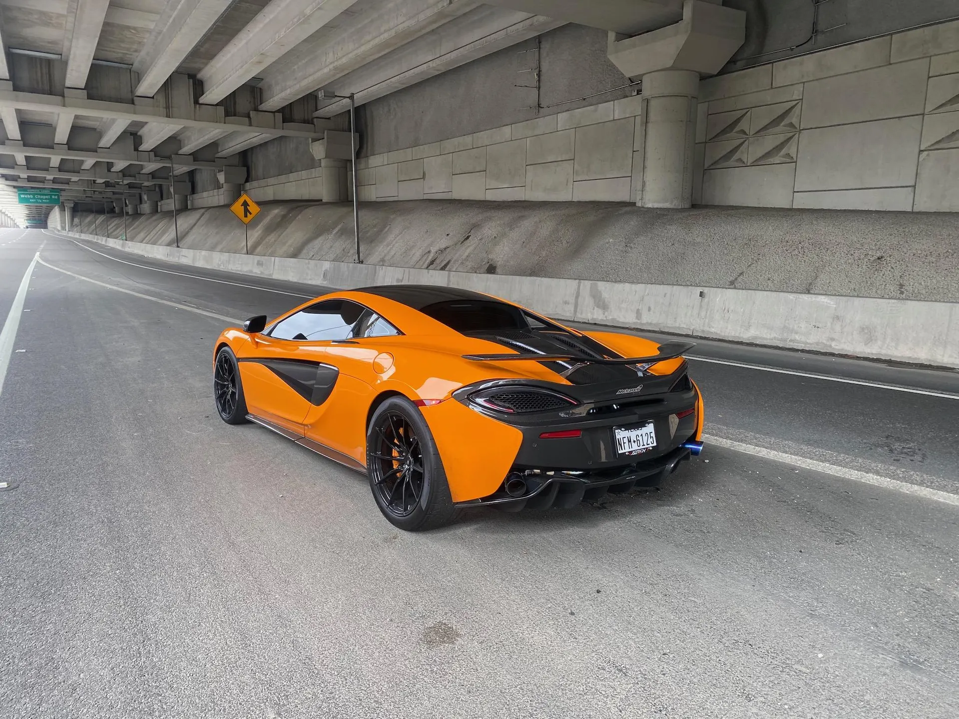 An orange McLaren 570S supercar parked on a road under a concrete overpass, showcasing its rear and side profile.