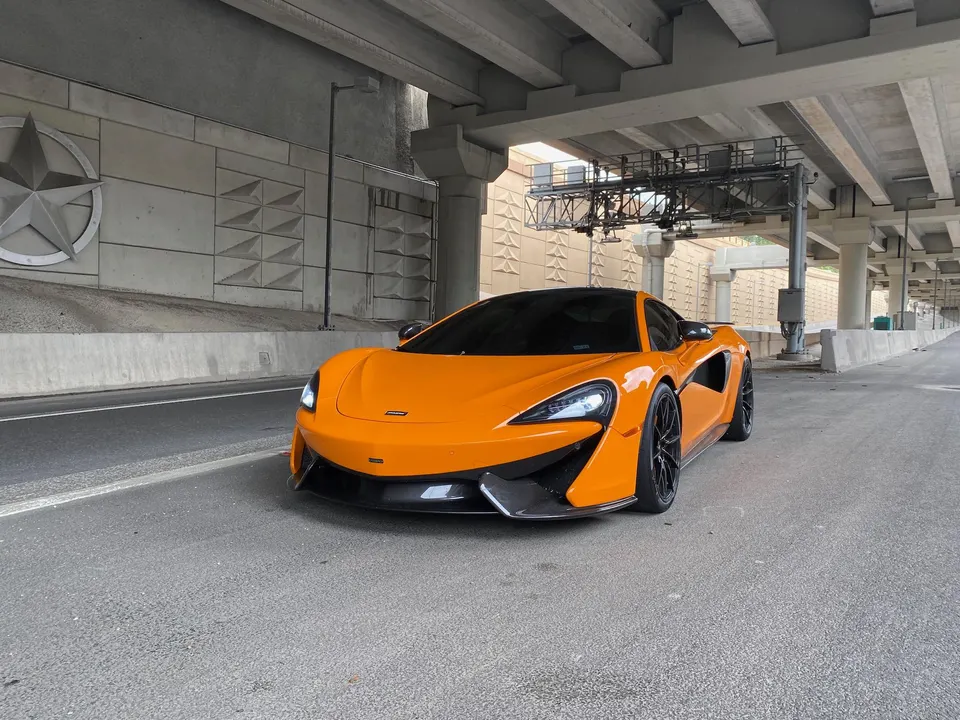 Orange McLaren supercar parked under a concrete overpass, showcasing its sleek design and carbon fiber accents.