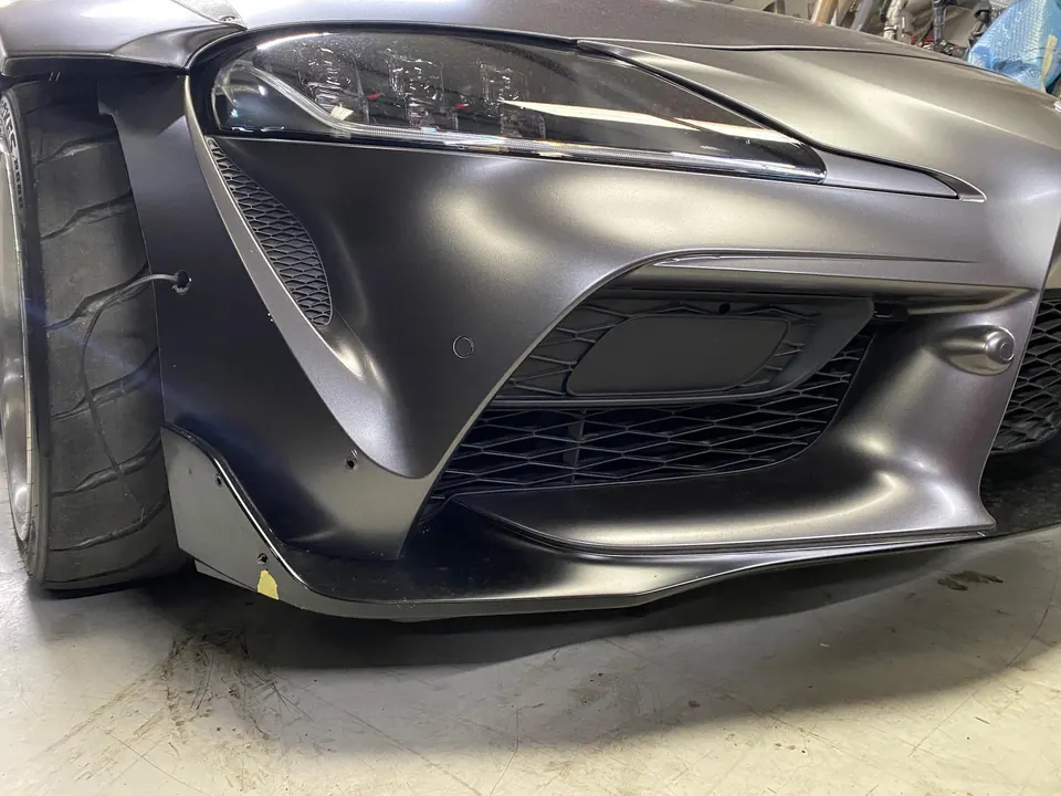 Matte grey Toyota Supra A90 with widebody fender, aggressive front splitter, and wide race tire in a shop setting.