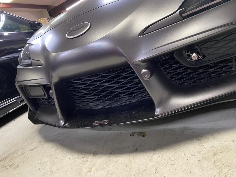 Close-up of a matte grey Toyota Supra A90 front bumper with a carbon fiber front lip and grille in a shop.