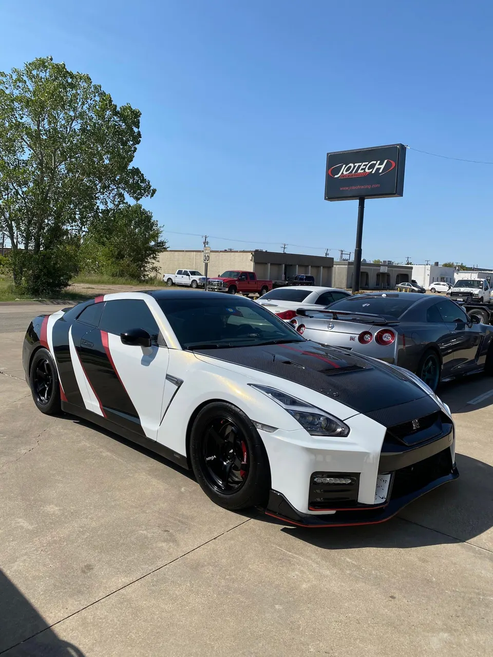 White, black, and red custom-wrapped Nissan GT-R R35 with a carbon fiber hood and aftermarket wheels parked at Jotech Motorsports.