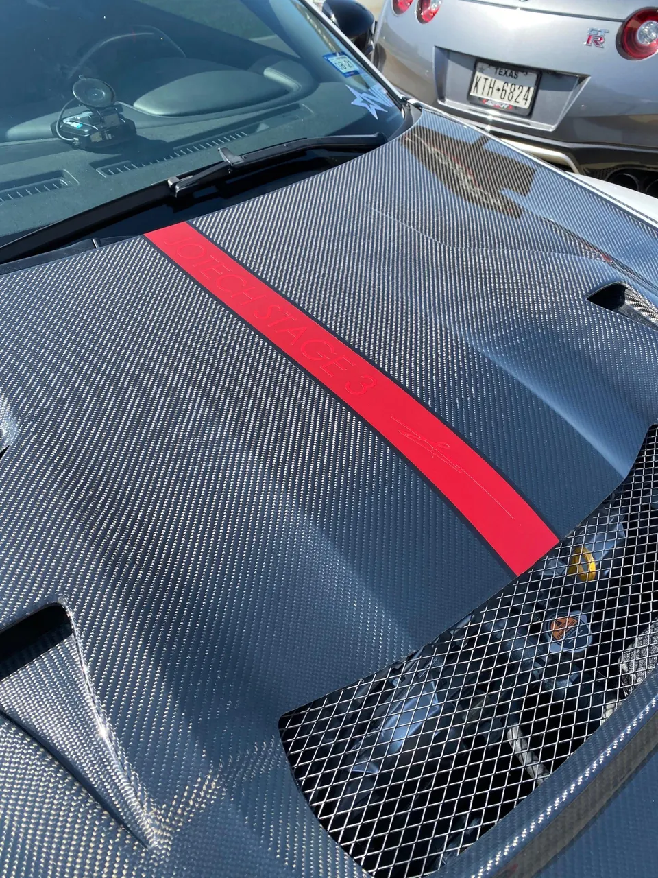 Close-up of a carbon fiber hood with a red "JOTECH STAGE 3" stripe and vents, with a Nissan GT-R in the background.